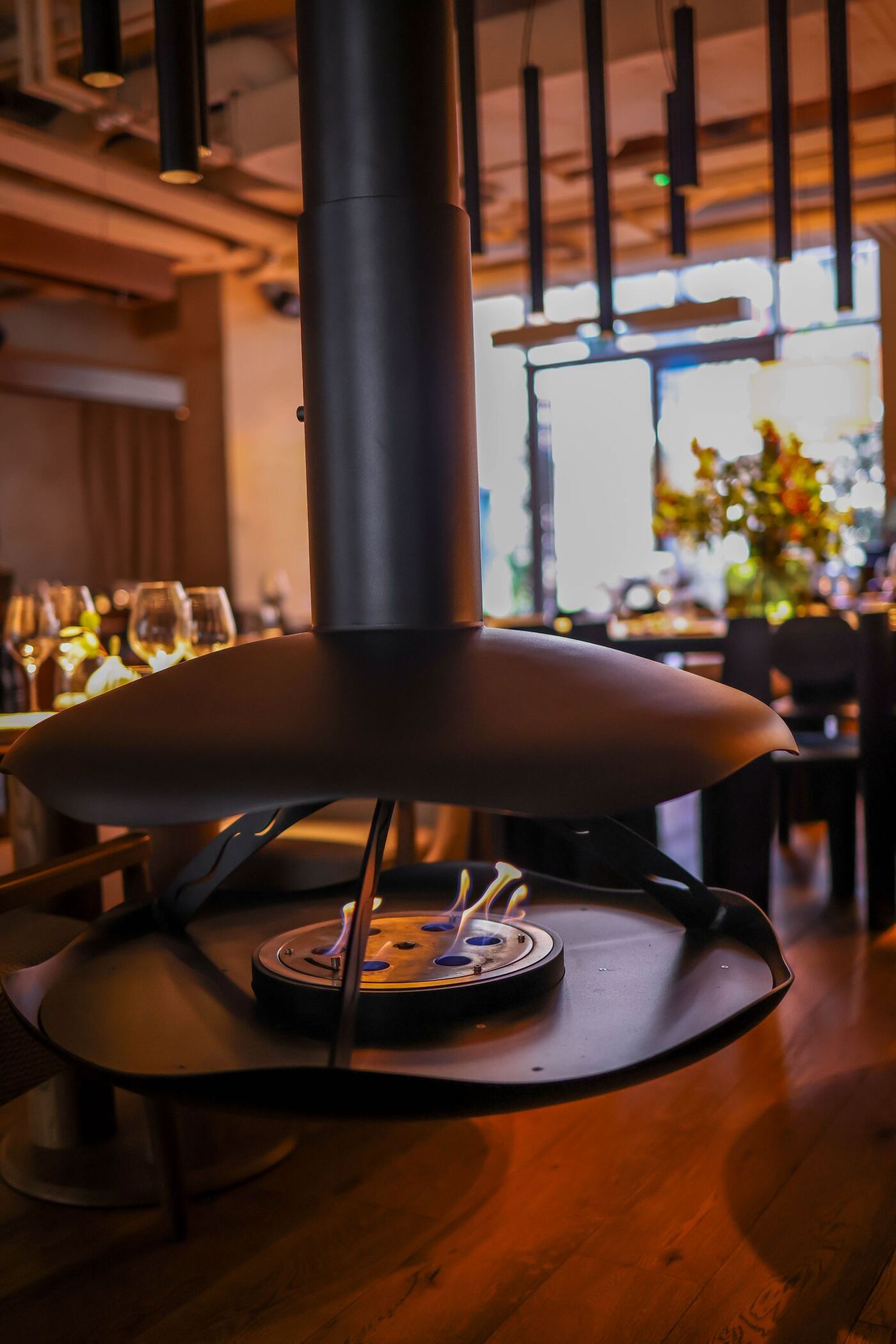 Comedor de madera con chimenea suspendida en el centro, mesas puestas y luz cálida de tarde